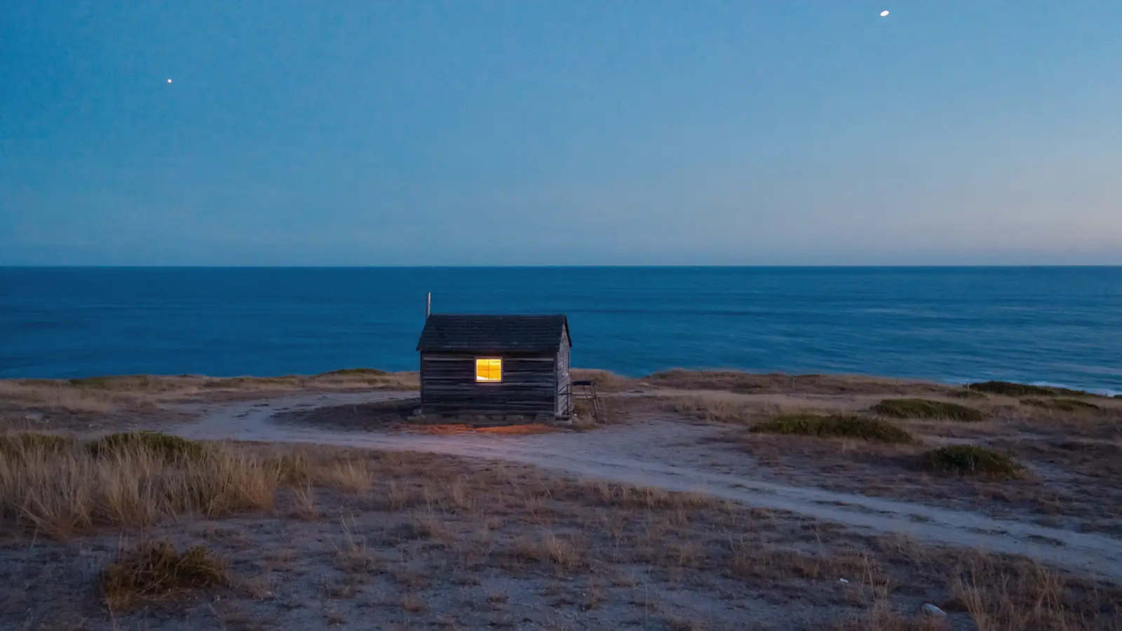 A small timber cabin alone at the end of a track on a headland at dusk, one gold window lit, the Pacific behind it