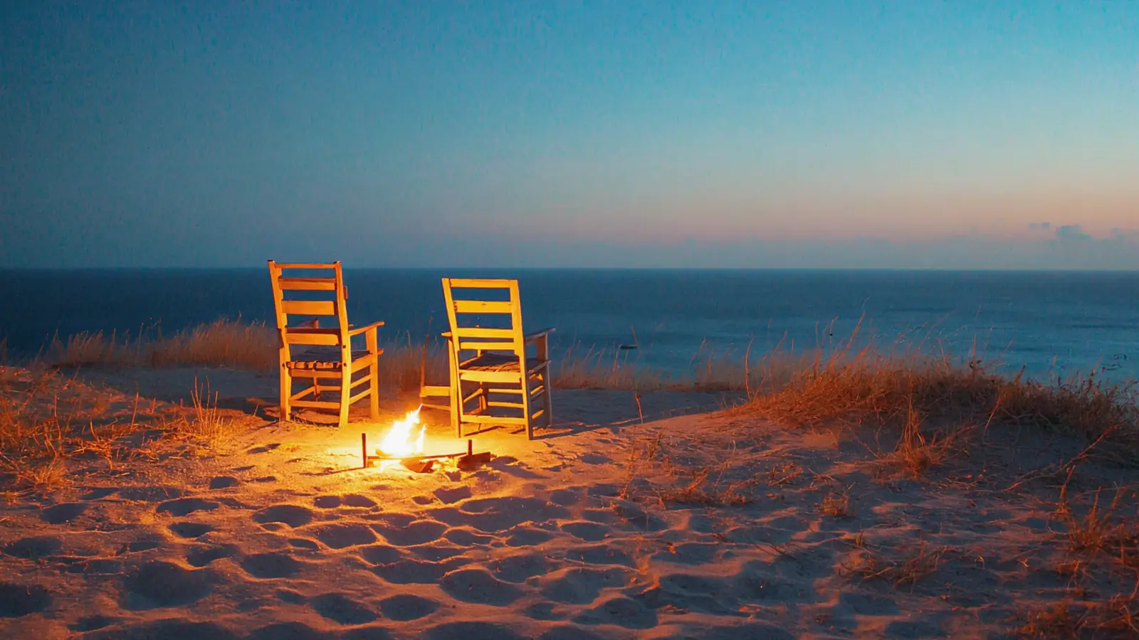 Two empty wooden chairs facing the sea at dusk, one of them lit by firelight