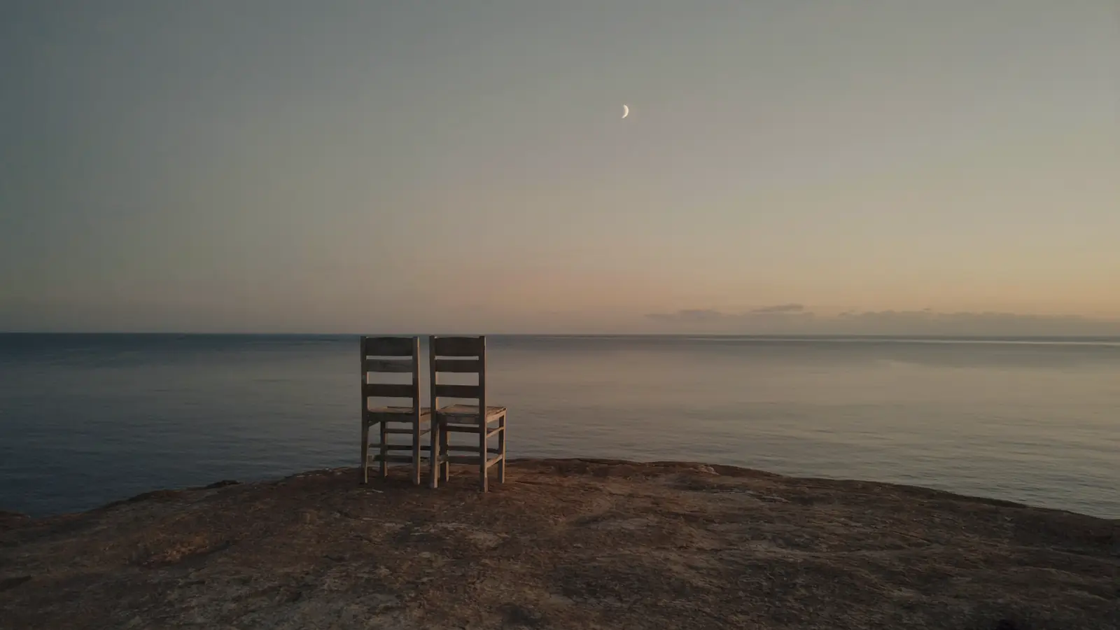 Two weathered chairs alone on bare rock, facing an empty sea